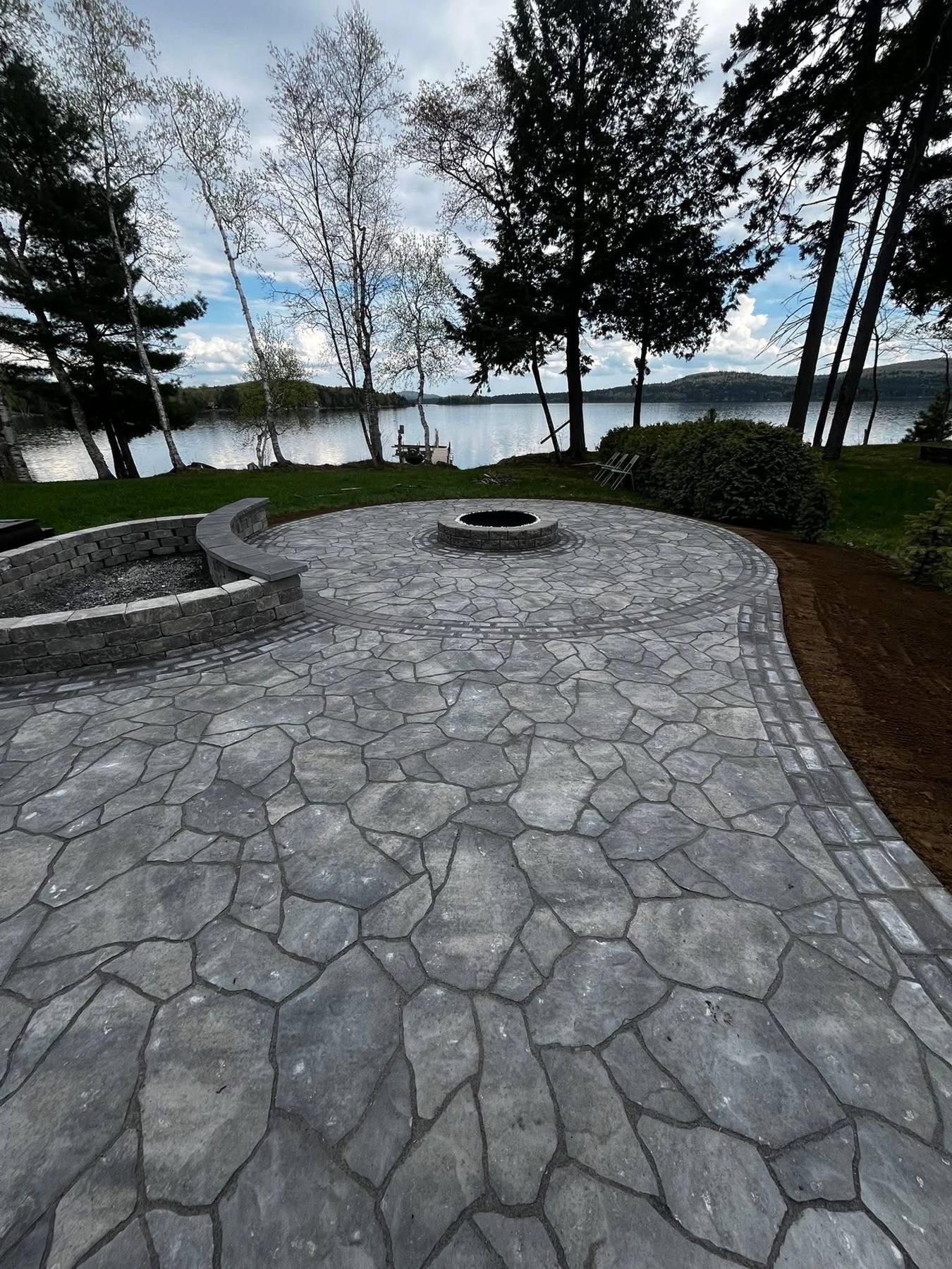 Grey flagstone patio with a round fire pit and curved seat wall overlooking a lake